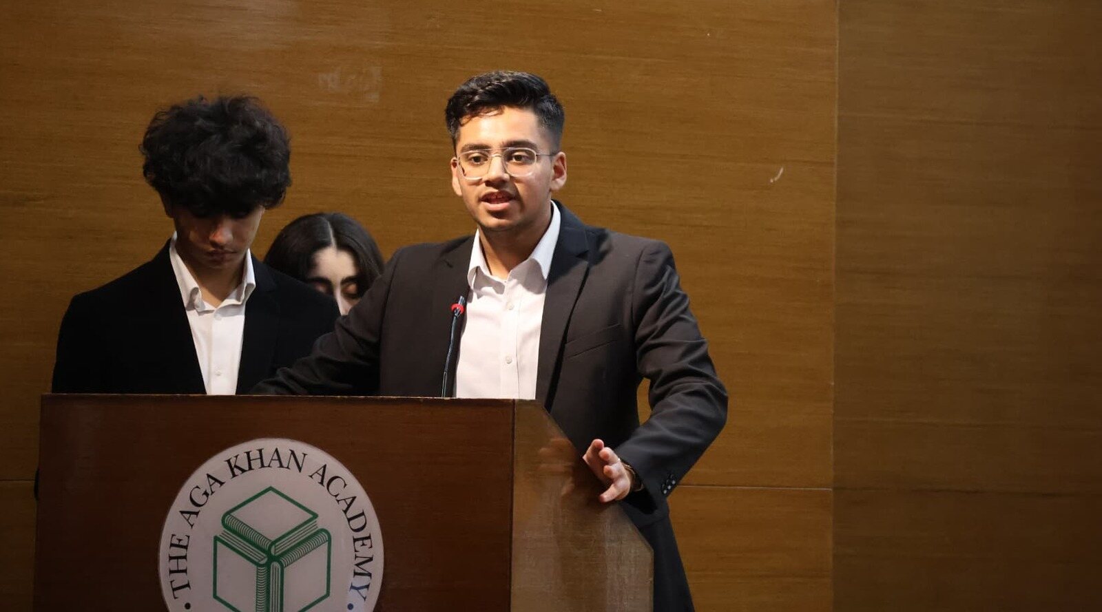 Emon Savani speaking at a podium bearing The Aga Khan Academy emblem